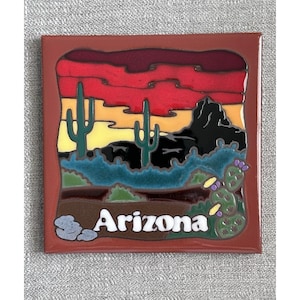 May include: A square ceramic tile featuring a desert landscape with two saguaro cacti, a black mountain silhouette, and a sunset sky in red, orange, and yellow. The word Arizona is printed in white letters at the bottom, with a prickly pear cactus.