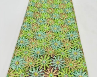 May include: A roll of lace fabric featuring a repeating pattern of embroidered flowers in shades of lime green, mint green, and soft gold. The floral design is connected by a fine, open-work mesh netting, creating a textured, semi-sheer textile.