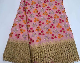 May include: Pink fabric with embroidered floral patterns in shades of pink, orange, and purple. The fabric has a gold lace trim along the bottom edge. The fabric is folded, showing the intricate details of the design.