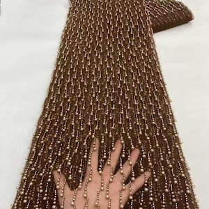 May include: Brown fabric with a beaded design. The fabric is covered in small, pearl-like beads that hang in vertical lines. The beads are a light, off-white color, creating a contrast with the brown background. The fabric is likely used for clothing or decorative purposes.
