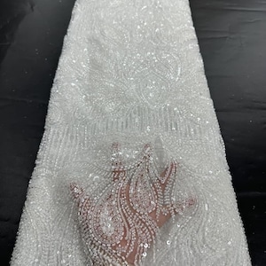 May include: A roll of white beaded fabric with intricate patterns. The fabric is heavily embellished with sequins and beads, creating a shimmering effect. The design features swirling, floral-like motifs. The fabric is suitable for bridal wear, evening gowns, or other special occasion garments.