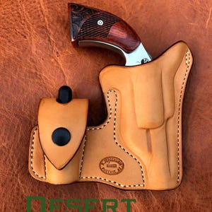 May include: A tan leather holster with a stitched design and a black button. The holster is designed to hold a small, silver revolver with a wood handle. The holster has a maker's mark that reads "R.J. Jorgenson Maker Phoenix, AZ".