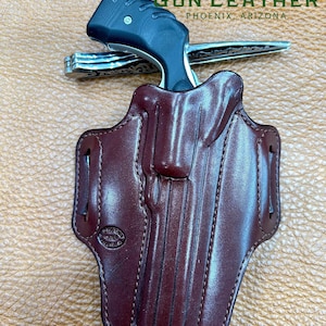 Puede incluir: Una funda de cuero marrón con un diseño cosido. La funda está diseñada para sostener un pequeño revólver. La funda está hecha por Desert Gun Leather, Phoenix, Arizona.