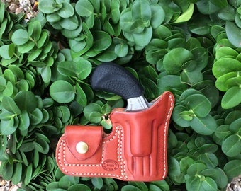 Könnte beinhalten: Ein braunes Lederholster mit einer kleinen schwarzen Pistole darin. Das Holster hat weiße Nähte und einen Messingknopf. Das Holster liegt auf einem Bett aus grünen Blättern.