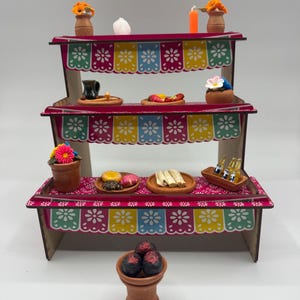 May include: A three-tiered wooden display stand decorated with colorful papel picado patterns in pink, yellow, blue, and green. The shelves hold miniature clay pots, food items, small bottles, and flowers. A separate clay bowl with dark items sits in front.