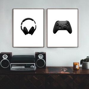 May include: Two framed prints on a white wall. The print on the left shows a pair of black headphones. The print on the right shows a black video game controller. The prints are hanging above a wooden shelf with a record player, two speakers, a candle, and a potted plant.