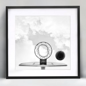 May include: A black and white photograph of a basketball hoop with a basketball going through the net. The hoop is against a cloudy sky.