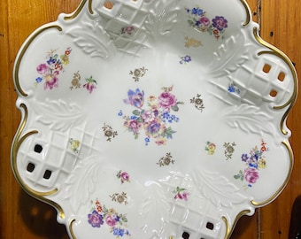 Puede incluir: Plato de porcelana blanca con borde festoneado y ribete dorado. El plato presenta un estampado floral en tonos morado, rosa, azul y amarillo. El diseño incluye secciones caladas y un motivo de hojas decorativas.