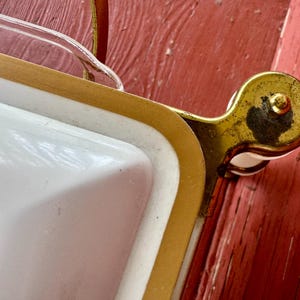 May include: Close-up of a vintage white serving tray with gold trim and a clear glass handle. The tray has a gold-colored metal handle and a white interior with a decorative design. The tray is on a red surface.