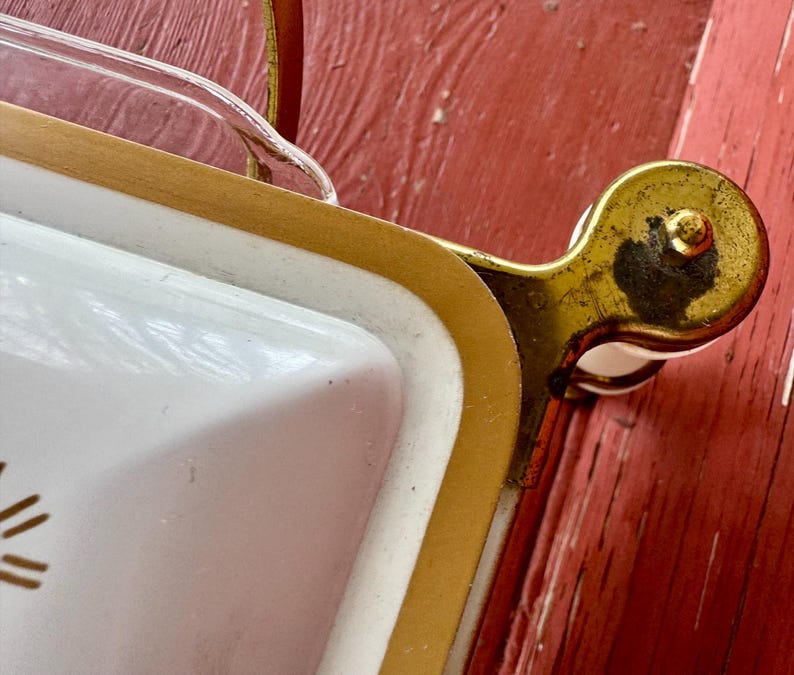 May include: Close-up of a vintage white serving tray with gold trim and a clear glass handle. The tray has a gold-colored metal handle and a white interior with a decorative design. The tray is on a red surface.