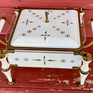 May include: A vintage white and gold serving dish with a square shape and a lid. The dish features a floral pattern and gold accents. It has white handles and legs with gold details. The dish is likely made of metal and glass.