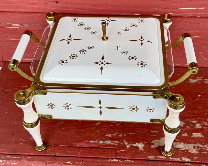 May include: A vintage white and gold serving dish with a square shape and a lid. The dish features a floral pattern and gold accents. It has white handles and legs with gold details. The dish is likely made of metal and glass.