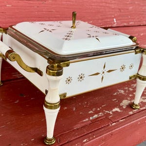 May include: White and gold serving dish with a lid, featuring a floral design. The dish has a rectangular shape, with a glass insert and is supported by four white legs with gold accents. The handles and lid handle are also gold.