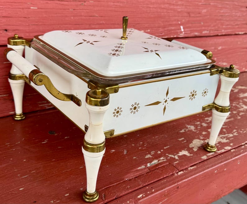 May include: White and gold serving dish with a lid, featuring a floral design. The dish has a rectangular shape, with a glass insert and is supported by four white legs with gold accents. The handles and lid handle are also gold.