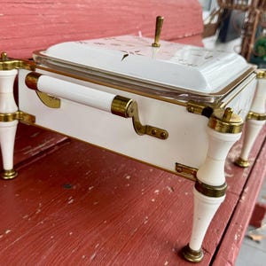 May include: A white and gold serving dish with a lid. The dish has a rectangular shape with rounded corners and is supported by four white legs with gold accents. The lid features a gold handle and a floral design.