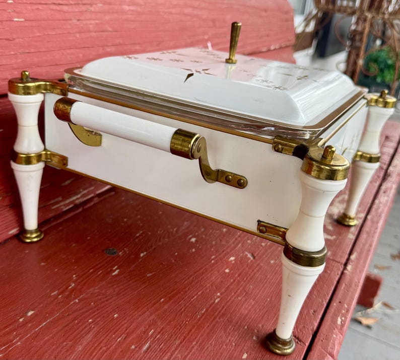 May include: A white and gold serving dish with a lid. The dish has a rectangular shape with rounded corners and is supported by four white legs with gold accents. The lid features a gold handle and a floral design.