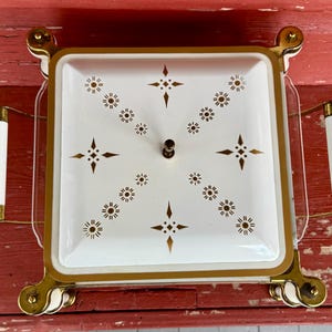 May include: A vintage square serving dish with a white interior and gold trim. The lid features a starburst pattern in gold. It has two white handles with gold accents and sits on gold-colored feet.