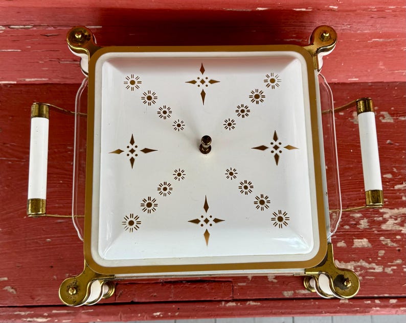 May include: A vintage square serving dish with a white interior and gold trim. The lid features a starburst pattern in gold. It has two white handles with gold accents and sits on gold-colored feet.