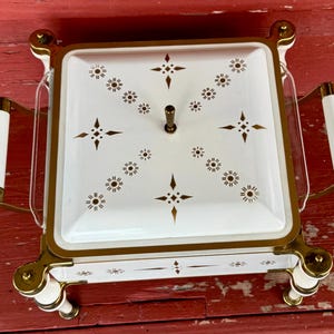 May include: A vintage white and gold serving dish with a square shape and a lid. The lid is decorated with a pattern of gold starbursts and dots. The dish has gold handles with white accents and sits on four white and gold feet.