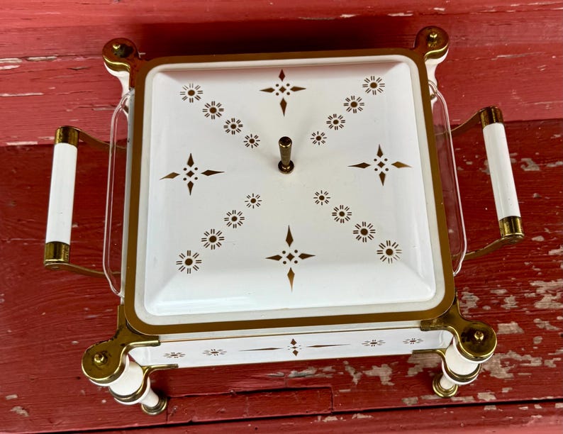May include: A vintage white and gold serving dish with a square shape and a lid. The lid is decorated with a pattern of gold starbursts and dots. The dish has gold handles with white accents and sits on four white and gold feet.