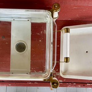May include: A vintage, square, glass and metal casserole dish with a lid. The dish has a clear glass body with a white interior and a white lid. It features gold-colored metal accents and white handles.