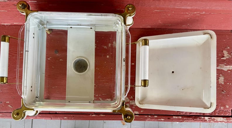 May include: A vintage, square, glass and metal casserole dish with a lid. The dish has a clear glass body with a white interior and a white lid. It features gold-colored metal accents and white handles.