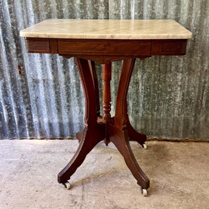 May include: A rectangular table featuring a smooth, cream-colored marble top set upon a dark brown wooden base. The table has four curved legs that converge at a central pillar and are equipped with small white casters for mobility.