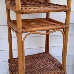 May include: A vintage, brown wicker plant stand with three shelves. The top shelf has a woven back and armrests. The middle shelf is slatted, and the bottom shelf is woven. The stand is made of natural materials.