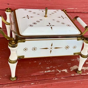 May include: A vintage white and gold serving dish with a lid. The dish features gold accents, decorative starburst and floral patterns, and white handles. The dish is elevated on four white legs with gold accents. A classic piece for entertaining.