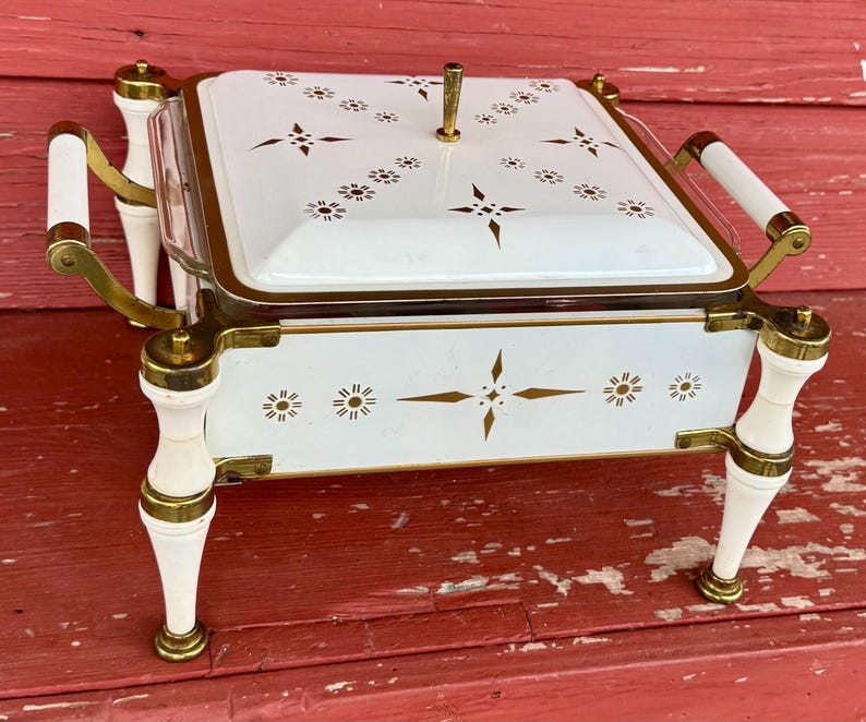 May include: A vintage white and gold serving dish with a lid. The dish features gold accents, decorative starburst and floral patterns, and white handles. The dish is elevated on four white legs with gold accents. A classic piece for entertaining.