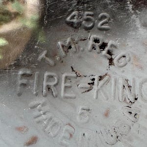 May include: Close-up of a gray, distressed metal surface with the embossed text "452", "T.M. REG", and "FIRE KING". The text "MADE IN USA" and the number "5" are also visible. The surface has scratches and wear.