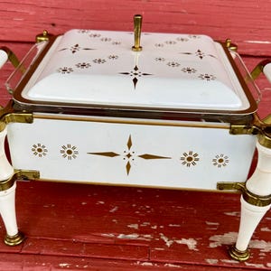 May include: A vintage white and gold serving dish with a lid. The dish features a rectangular shape with a clear glass insert, gold accents, and white handles. The lid and base are decorated with gold starburst and geometric patterns.