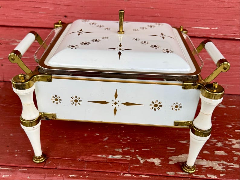 May include: A vintage white and gold serving dish with a lid. The dish features a rectangular shape with a clear glass insert, gold accents, and white handles. The lid and base are decorated with gold starburst and geometric patterns.
