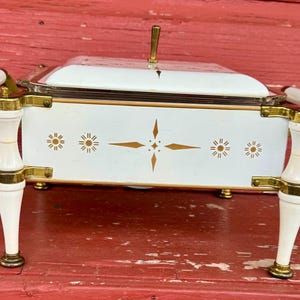 May include: A white and gold vintage serving dish with a clear glass lid. The dish has gold accents, decorative starburst designs, and white handles. The dish is on four white and gold legs.