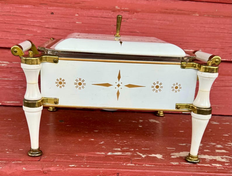 May include: A white and gold vintage serving dish with a clear glass lid. The dish has gold accents, decorative starburst designs, and white handles. The dish is on four white and gold legs.