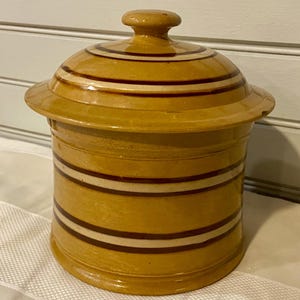 May include: A yellow ceramic jar with a lid. The jar has horizontal bands of white and brown stripes. The lid has a small knob on top. The jar is likely used for storage.