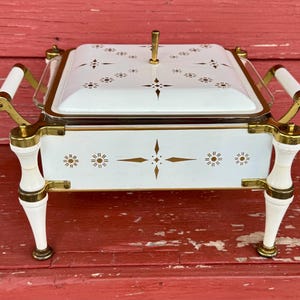 May include: A white and gold serving dish with a lid, featuring a decorative floral pattern. The dish has gold handles and legs, and a gold accent around the rim. The dish is on a red wooden surface.