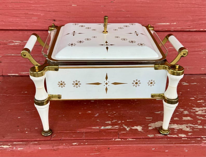 May include: A white and gold serving dish with a lid, featuring a decorative floral pattern. The dish has gold handles and legs, and a gold accent around the rim. The dish is on a red wooden surface.