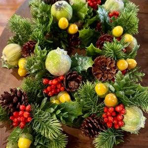 May include: A festive Christmas wreath featuring green holly leaves, pine needles, and brown pine cones. The wreath is adorned with yellow berries, green apples, and red berries. A gold-colored key with the text "MARY DNA" is also present.