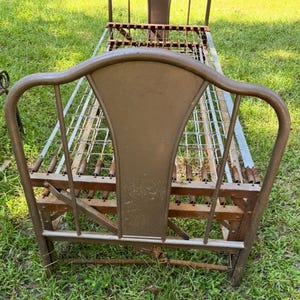 May include: An antique metal bed frame with a headboard and spring mattress. The frame is a muted bronze color, showing signs of age and wear. The headboard has a curved top and a central panel. The bed frame is set on a grassy surface.