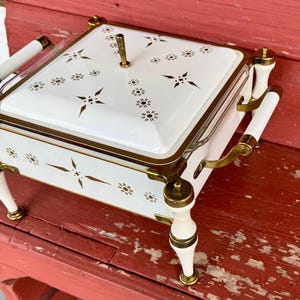 May include: A vintage white and gold serving dish with a lid. The dish features a square shape with a gold trim and a handle on top. The lid and sides are decorated with gold starburst and floral patterns. The dish has white handles and legs with gold accents.
