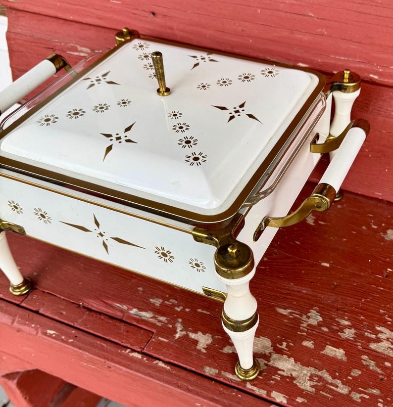 May include: A vintage white and gold serving dish with a lid. The dish features a square shape with a gold trim and a handle on top. The lid and sides are decorated with gold starburst and floral patterns. The dish has white handles and legs with gold accents.
