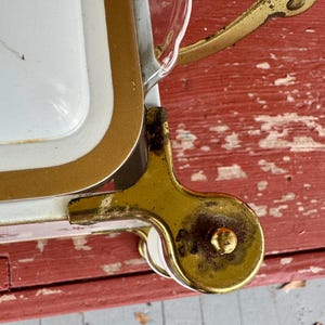 May include: Close-up of a vintage white and gold serving tray with a gold handle and a decorative wheel. The tray has a white interior with a gold border. The handle and wheel are made of gold-colored metal.