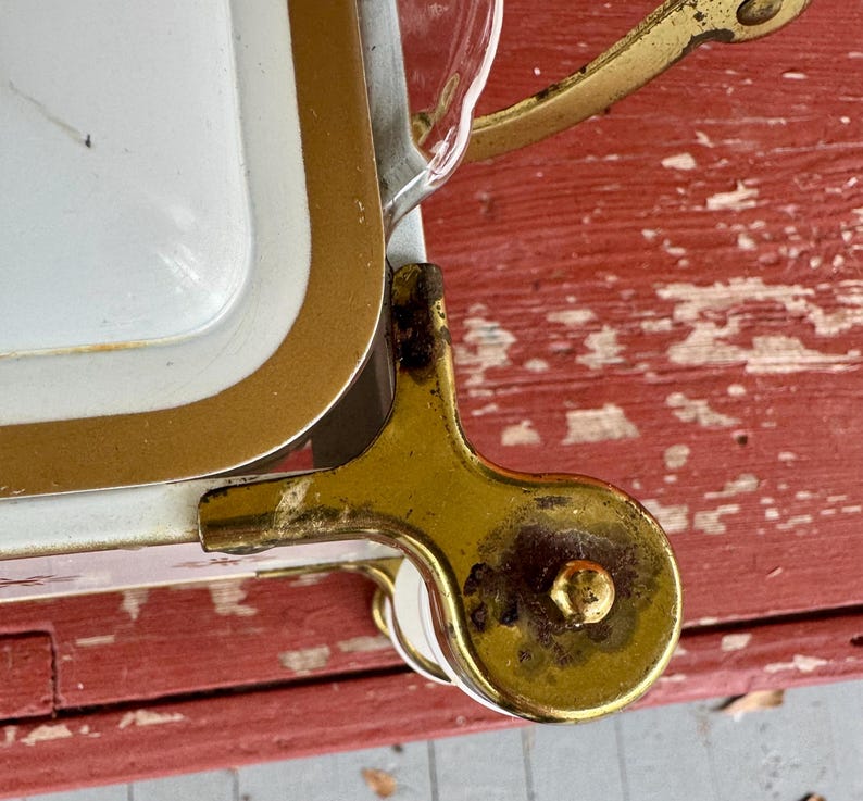 May include: Close-up of a vintage white and gold serving tray with a gold handle and a decorative wheel. The tray has a white interior with a gold border. The handle and wheel are made of gold-colored metal.