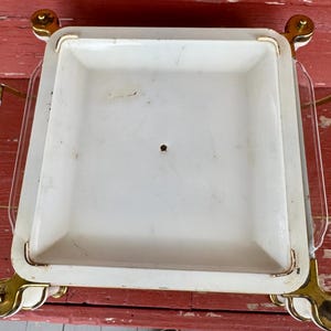 May include: A vintage serving tray with a white square dish and clear glass sides. The tray has gold-colored accents on the corners and handles, and white cylindrical handles. The tray is on a red surface.