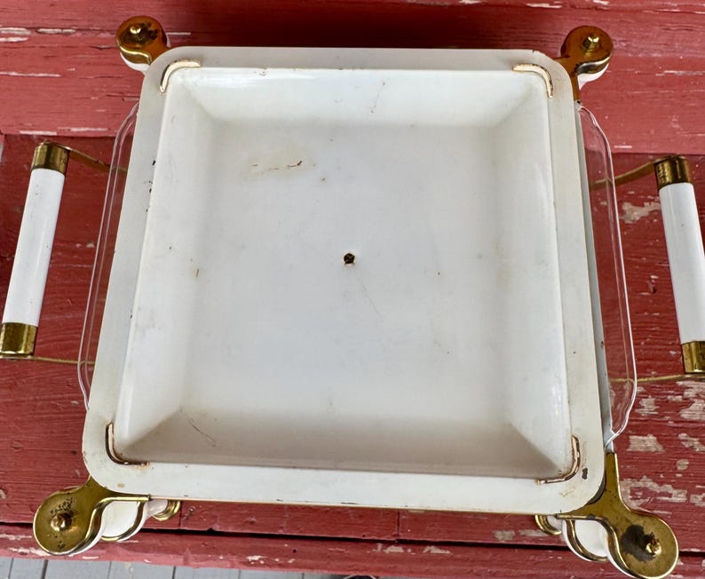 May include: A vintage serving tray with a white square dish and clear glass sides. The tray has gold-colored accents on the corners and handles, and white cylindrical handles. The tray is on a red surface.