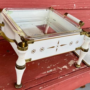 May include: A vintage white and gold serving dish with a clear glass insert. The dish has decorative gold accents, white handles, and a matching lid. The design includes starburst and floral patterns, perfect for serving food.