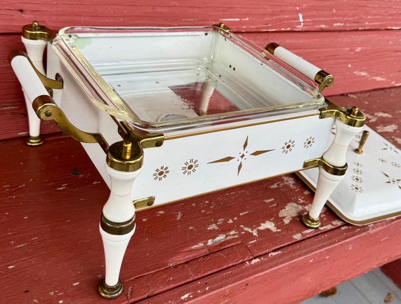 May include: A vintage white and gold serving dish with a clear glass insert. The dish has decorative gold accents, white handles, and a matching lid. The design includes starburst and floral patterns, perfect for serving food.