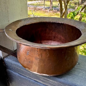 May include: A large, antique copper chamber pot with a wide, rounded rim and a single handle. The pot has a rich, reddish-brown patina, showing signs of age and use. The interior is dark, and the exterior has a hammered texture.