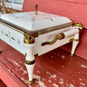 May include: White and gold vintage serving dish with a glass lid. The dish has gold accents, a handle, and decorative starburst and floral patterns. The legs and handles are white with gold trim. A gold knob is on the lid.